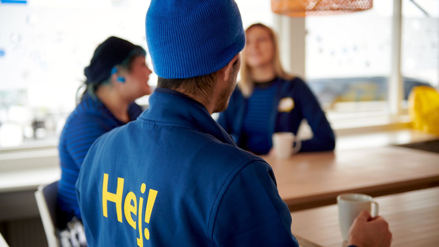 Picture of three employees inside a lunch room, closest to me sits a guy wearing blue Ikea clothes and a blue cap with the text HEJ on the back.