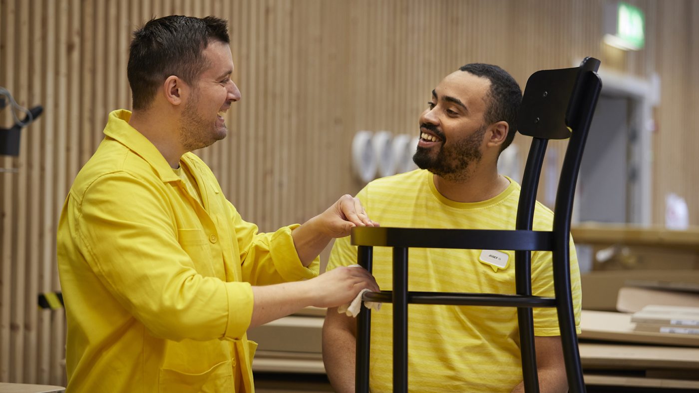 Two IKEA colleagues are talking to each other and laughing with a chair in front of them that they are holding.