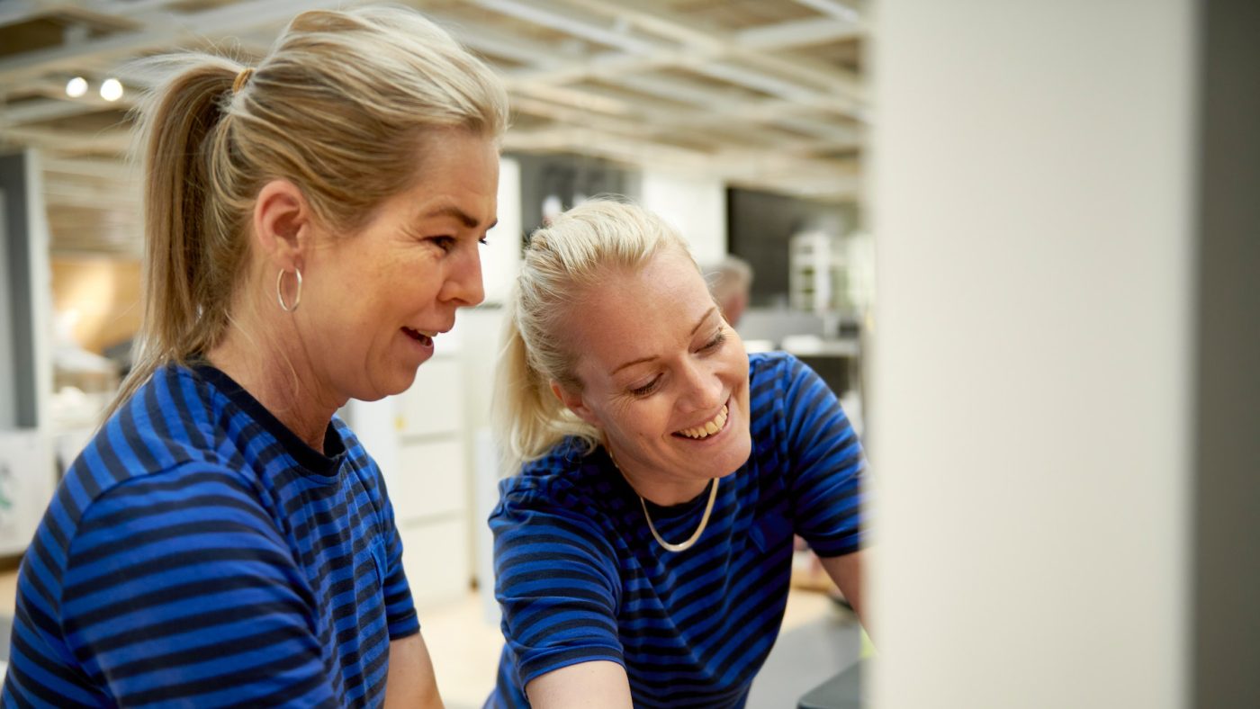 Two female colleagues laugh with room numbers while fixing an IKEA product inside a department store.