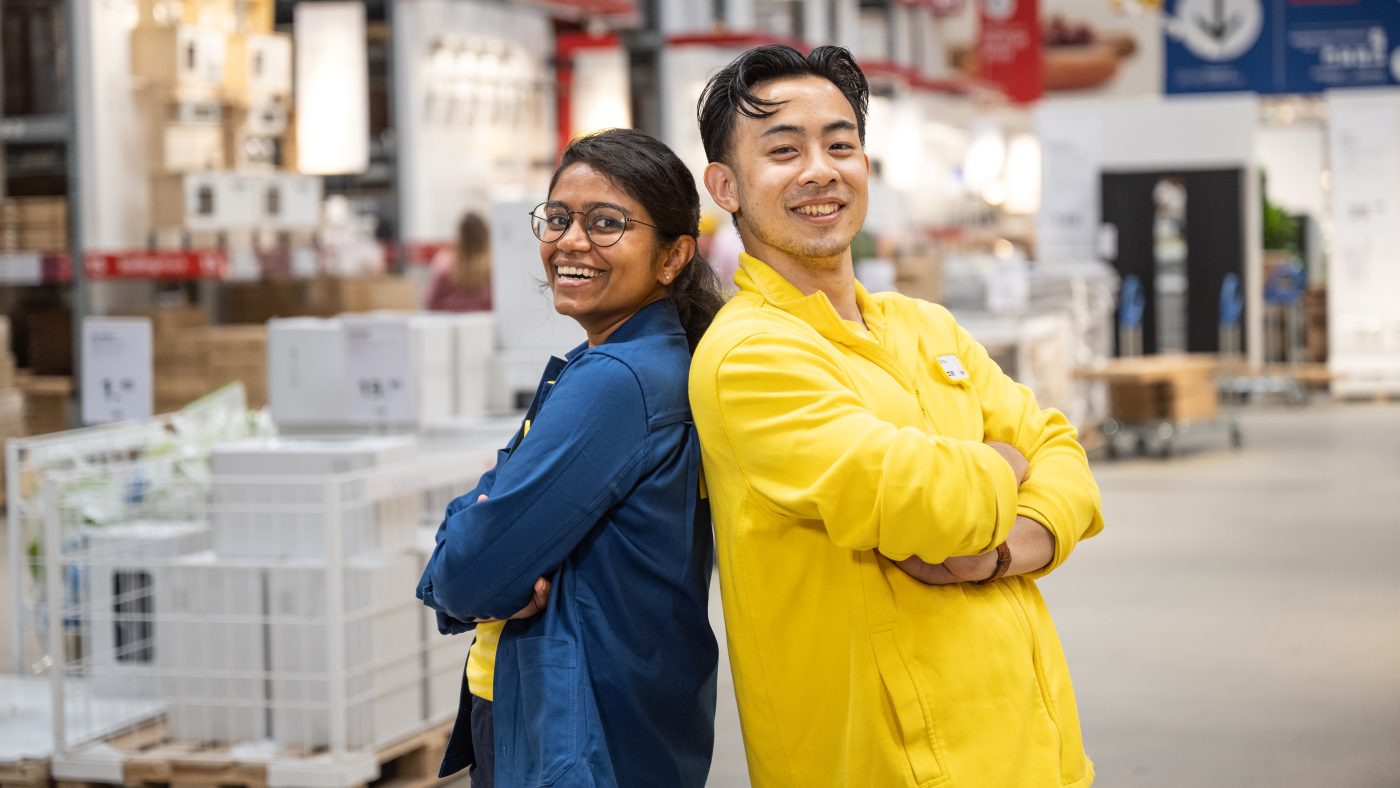 Two colleagues, a girl and a guy stand with their backs to each other and smile at the camera in yellow and blue clothes inside an IKEA store.