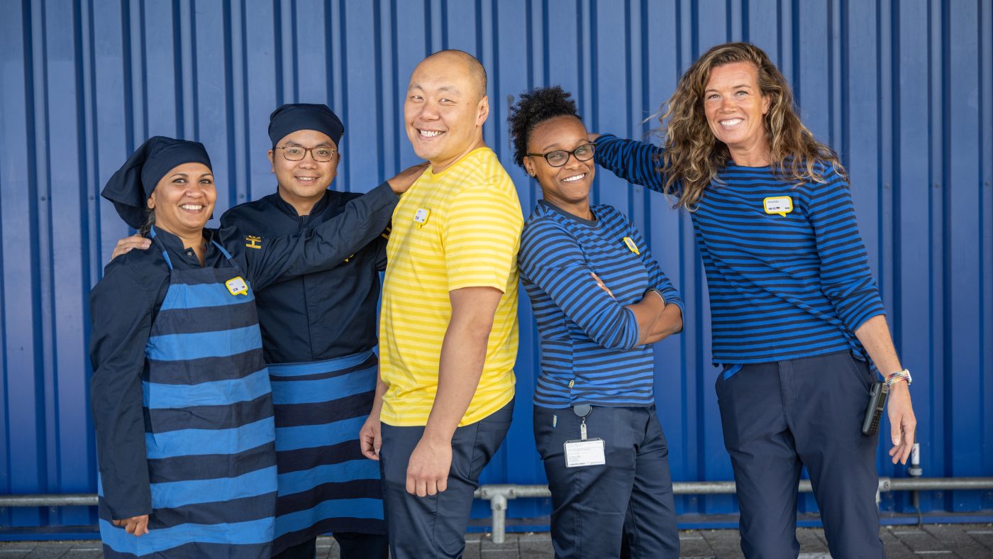 Picture of many IKEA colleagues outside an IKEA department store, laughing and smiling at the camera wearing IKEA work clothes.