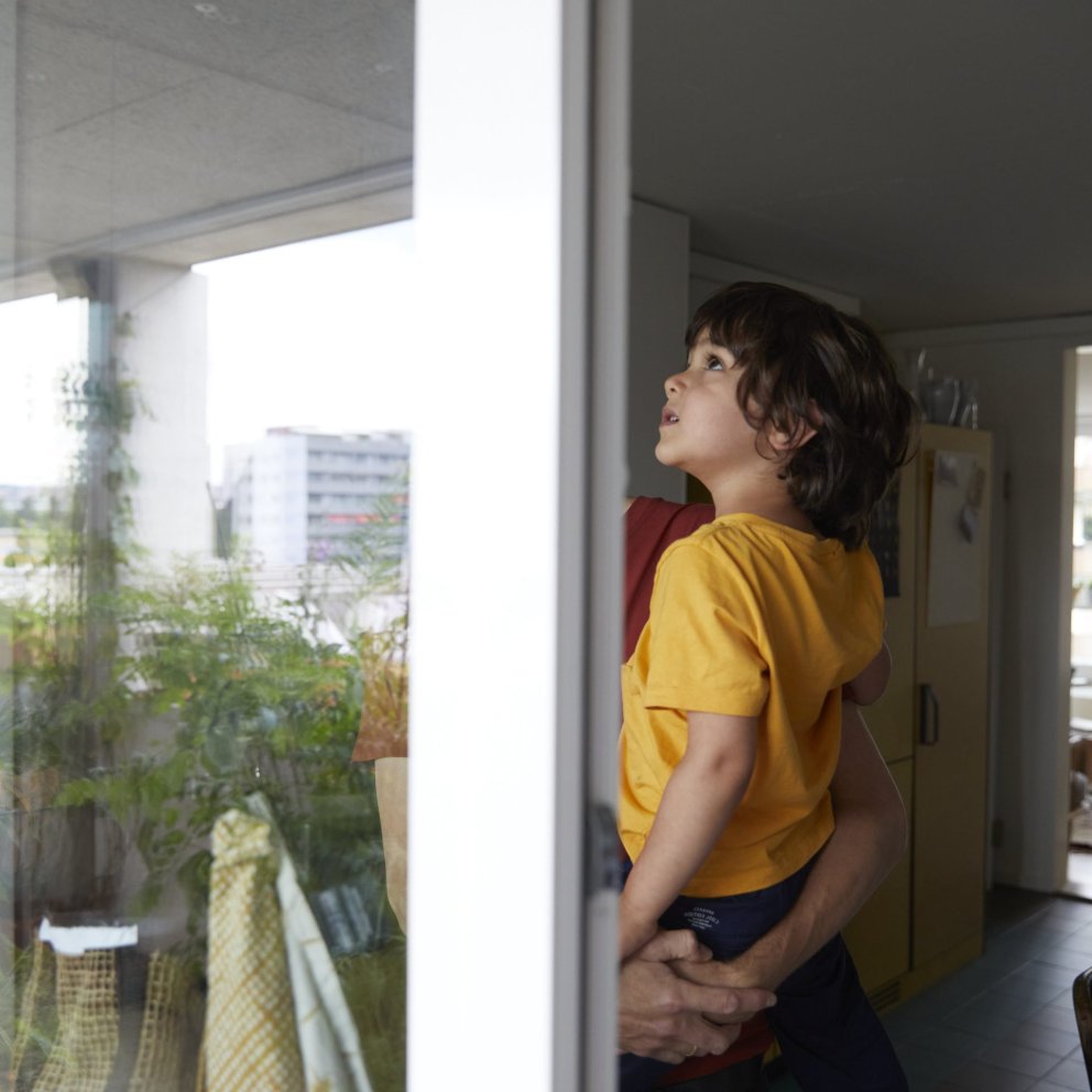 A man holds his child in his arms and looks out of a window with the living room in the background.