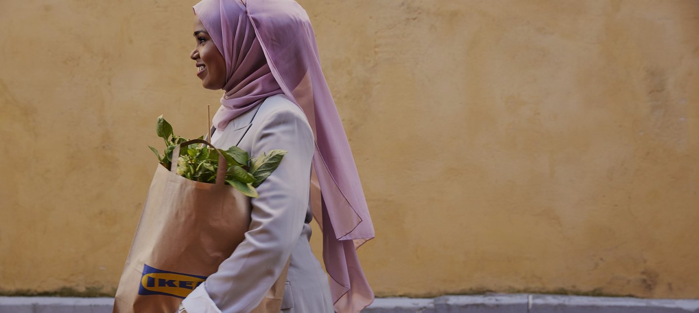 A girl walks with an IKEA bag of plants in her hand where the picture is taken in profile against a yellow wall.