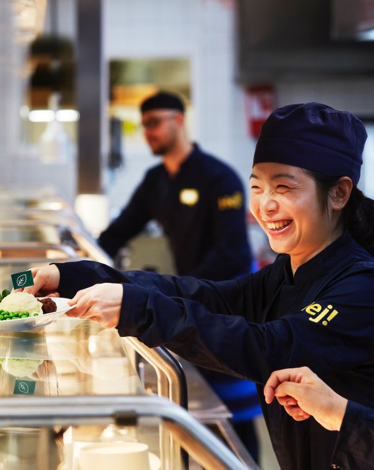 Three employees in the IKEA kitchen where one laughs and hands out food to customers in the restaurant.