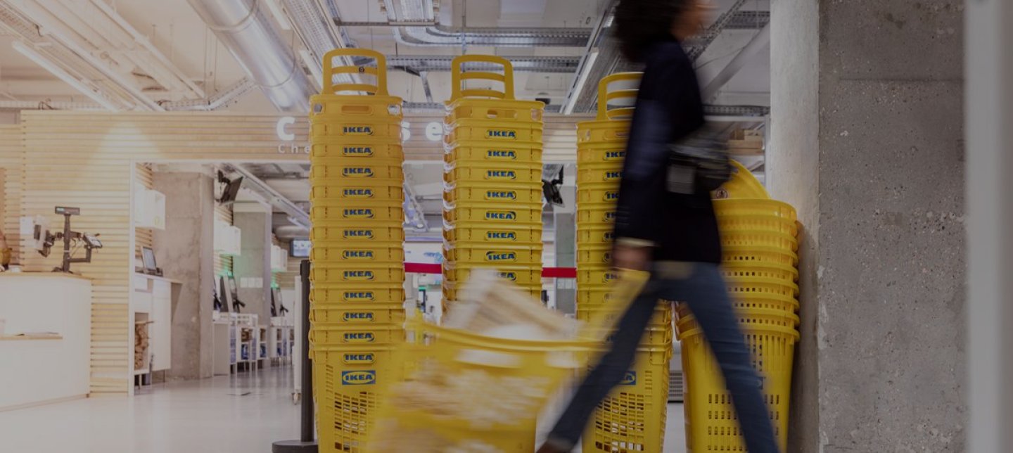Girl walks inside an IKEA department store with a shopping basket and the background is filled with shopping baskets. The picture is also taken in profile.